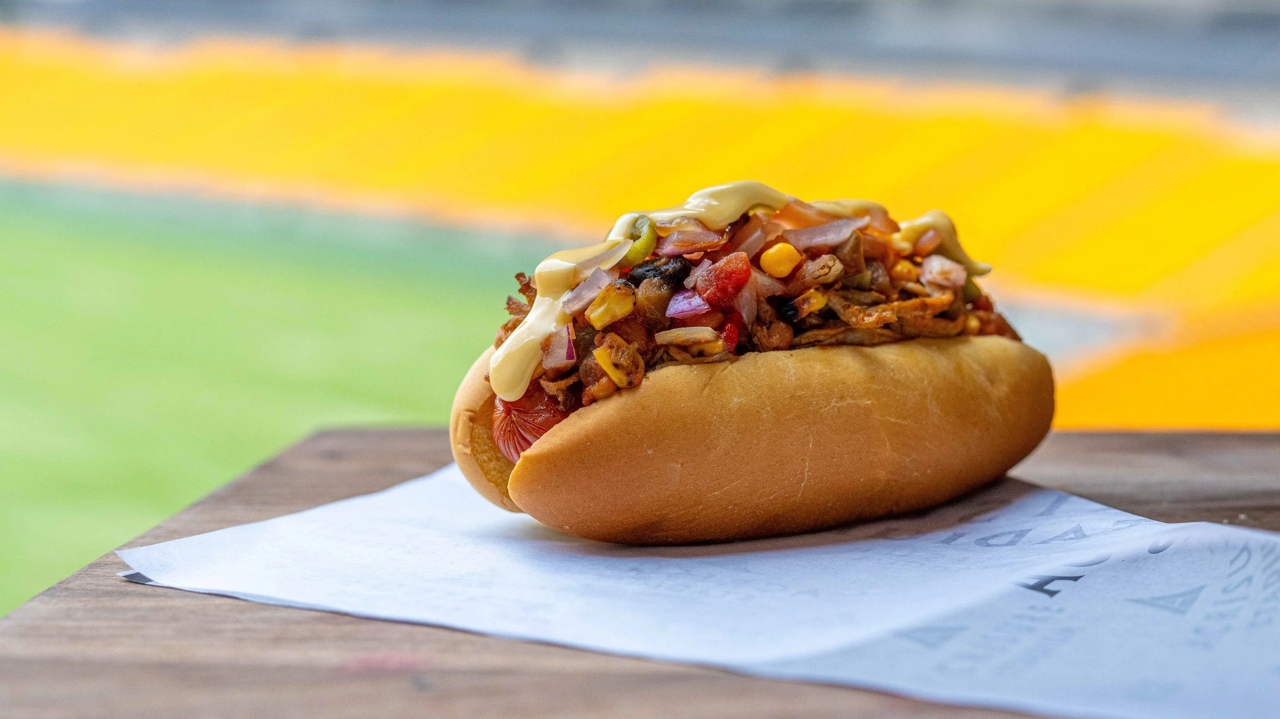 Loaded stadium hot dog topped with shredded meat, corn, black beans, diced peppers and creamy sauce on Acrisure Stadium-branded paper, photographed by Ian Jones of ISJDESIGNS in Pittsburgh for Aramark Sports + Entertainment, an ISJDESIGNS food photography client.