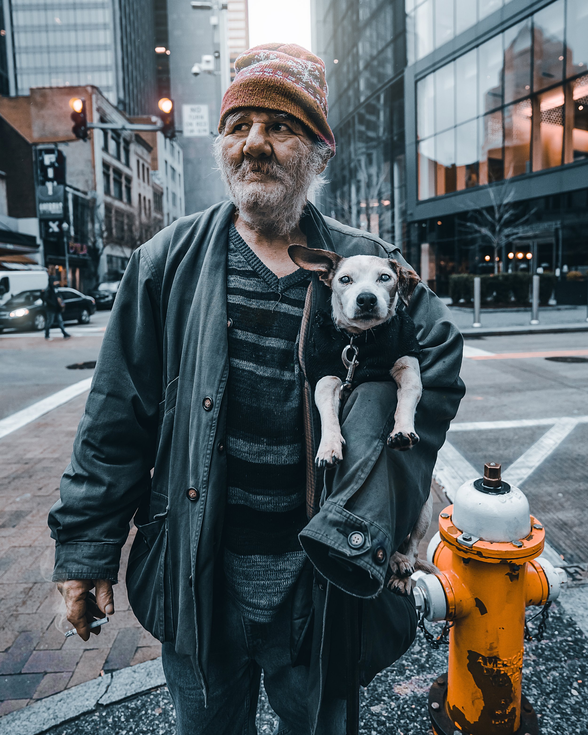 Street portrait by Ian Jones of an older man holding a small dog beside an orange fire hydrant in an urban downtown setting for a Photofocus Tamron Recipes feature.