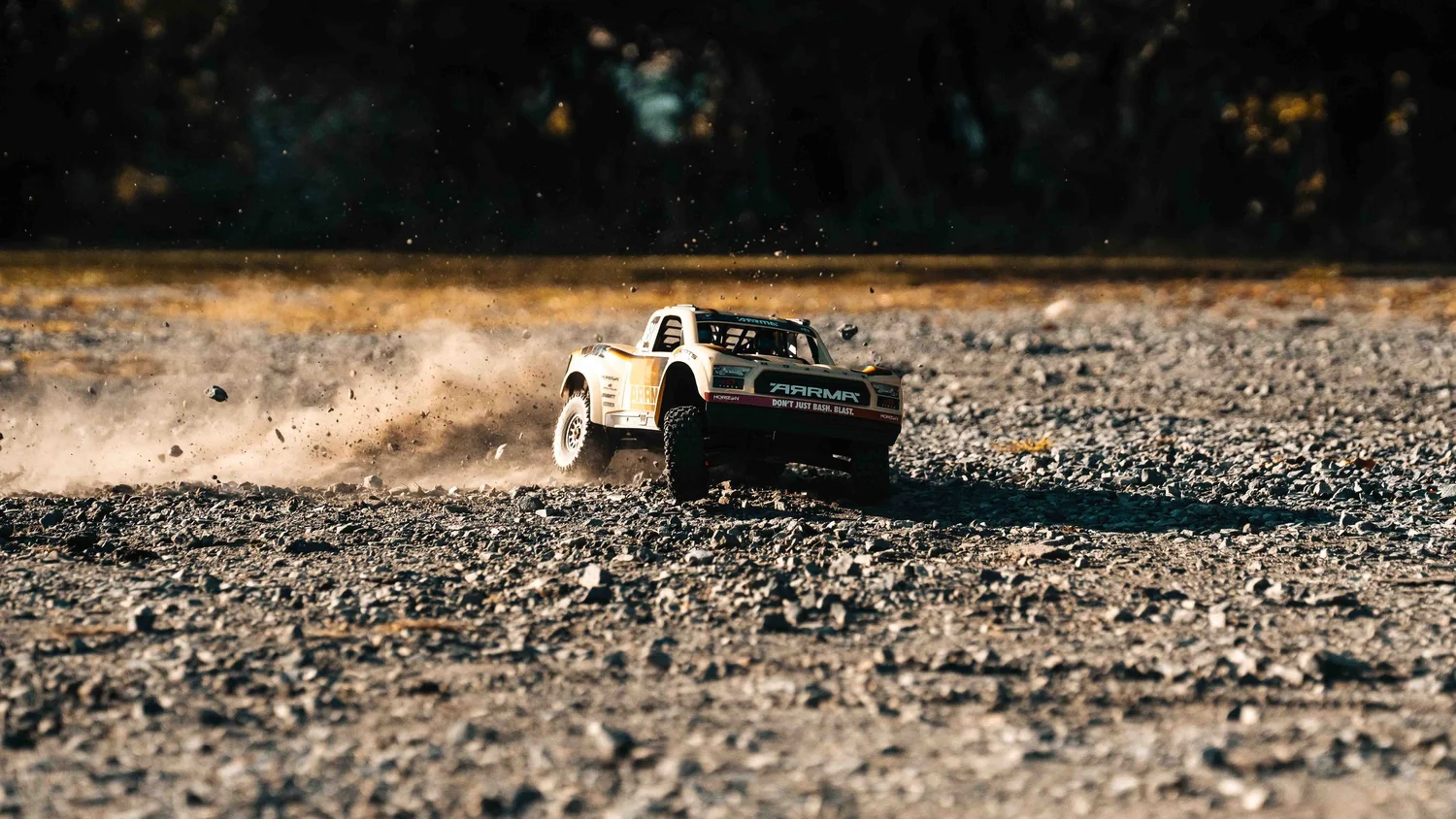 Wide environmental photograph of the ARRMA Mojave 6S V3 RC truck driving across an open rocky clearing with dark trees in the background.