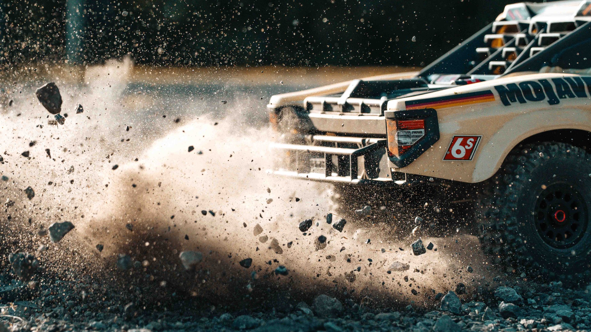 Dramatic close-up of the ARRMA Mojave 6S V3 desert truck throwing gravel and dust from its front wheel, captured in warm sunlight with individual stones frozen in motion.