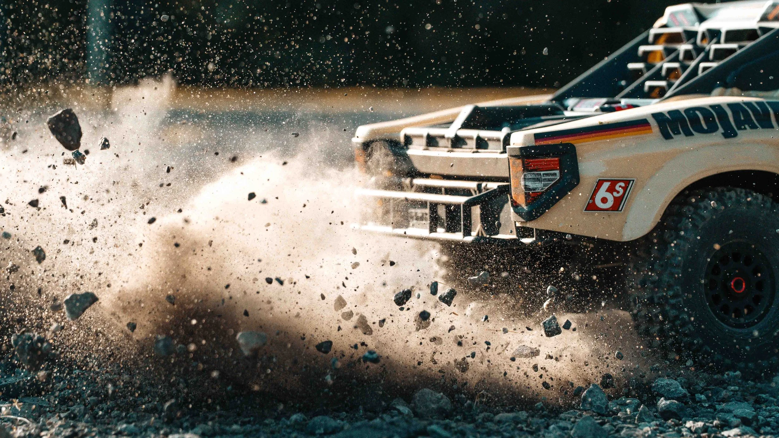 Dramatic close-up of the ARRMA Mojave 6S V3 desert truck throwing gravel and dust from its front wheel, captured in warm sunlight with individual stones frozen in motion.