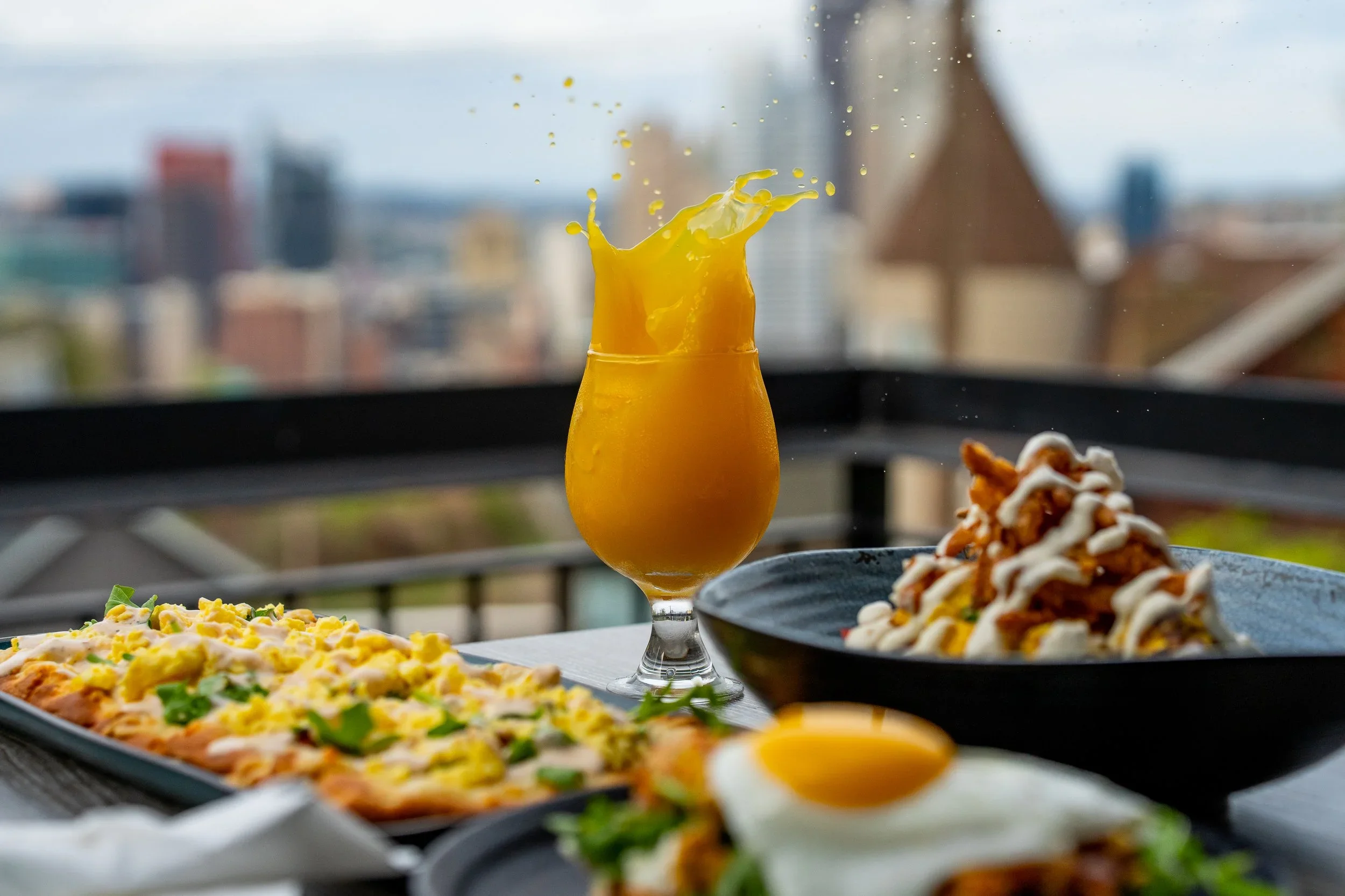 Rooftop food photography by Ian Jones featuring an orange drink splash, plated food, and a blurred city skyline for a ProMaster Photo feature.