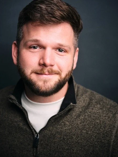 Dennis Connors headshot, theatrical: soft smile, olive quarter-zip, dark backdrop.