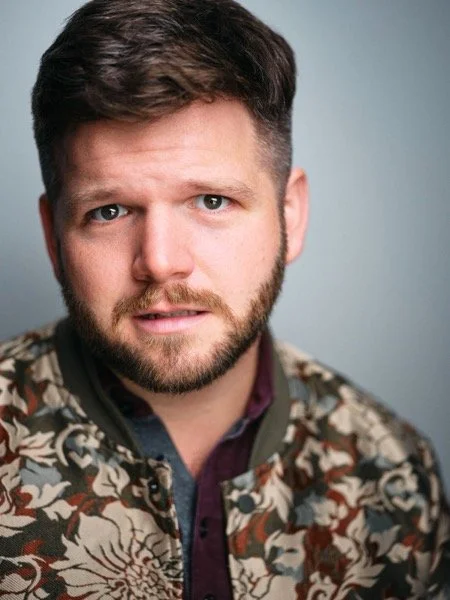 Dennis Connors headshot, drama: direct unsmiling look to camera, floral bomber jacket, grey backdrop.