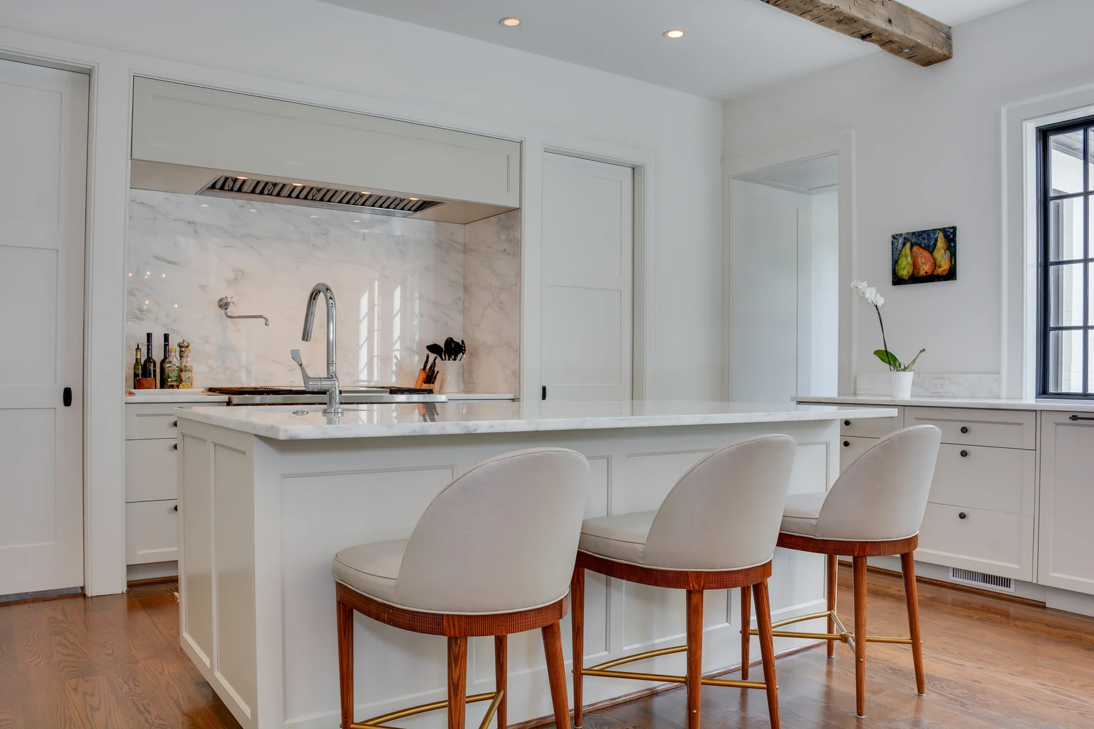 Sunlit white kitchen with upholstered stools, warm wood flooring, and a beamed ceiling