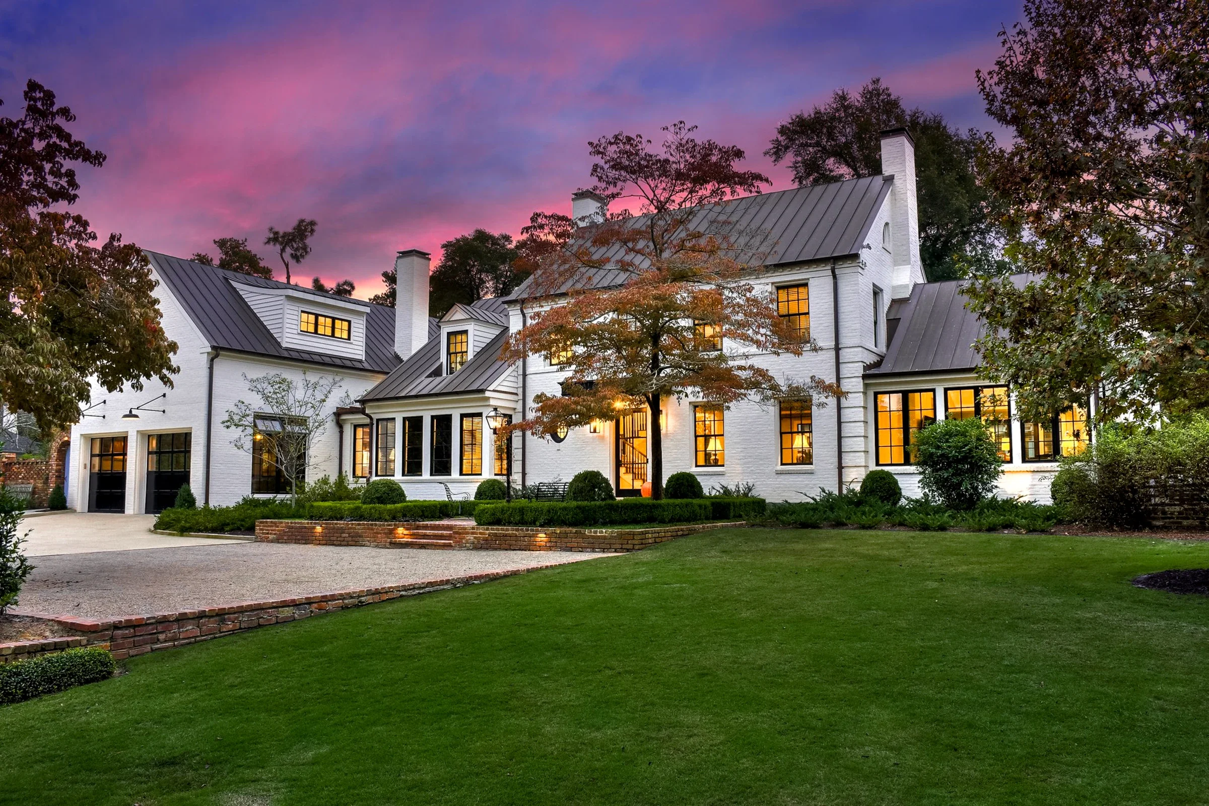 A white brick home with illuminated windows and a broad green lawn beneath a pink twilight sky