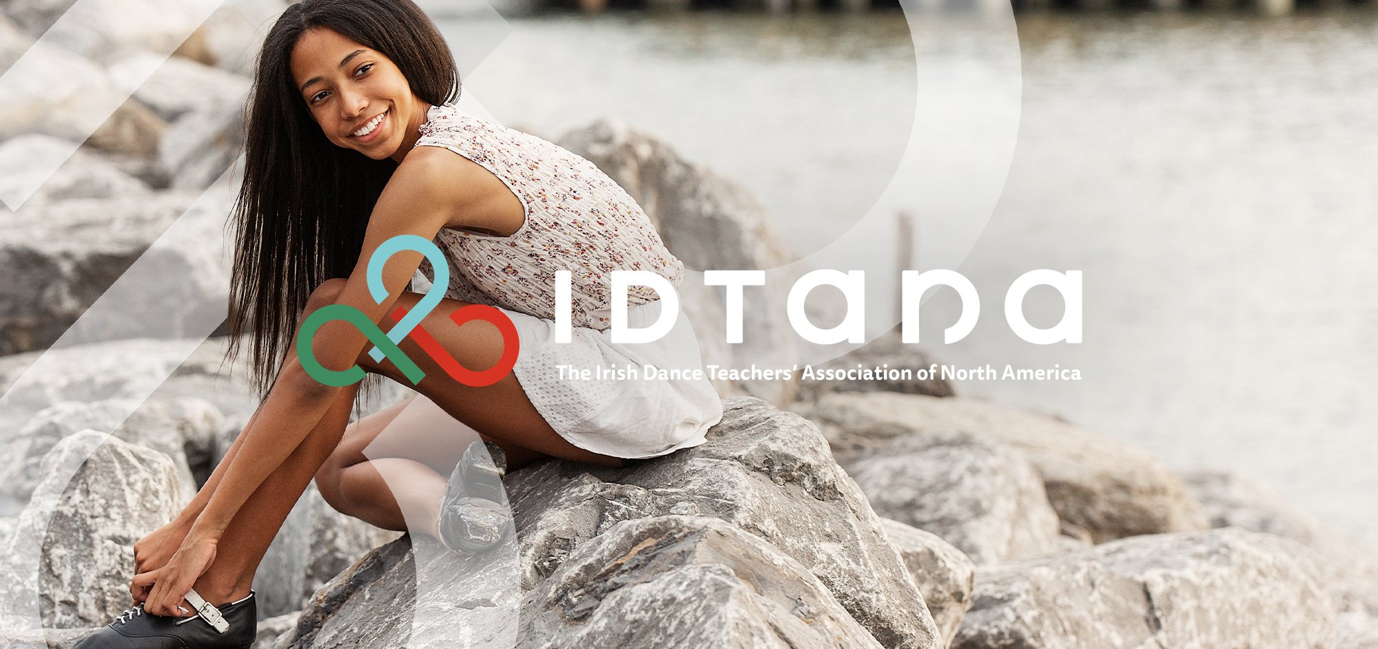 A picture of a girl tying her Irish dance shoes while sitting on rocks near the East River in New York. There is an overlay of the IDTANA logo on top.