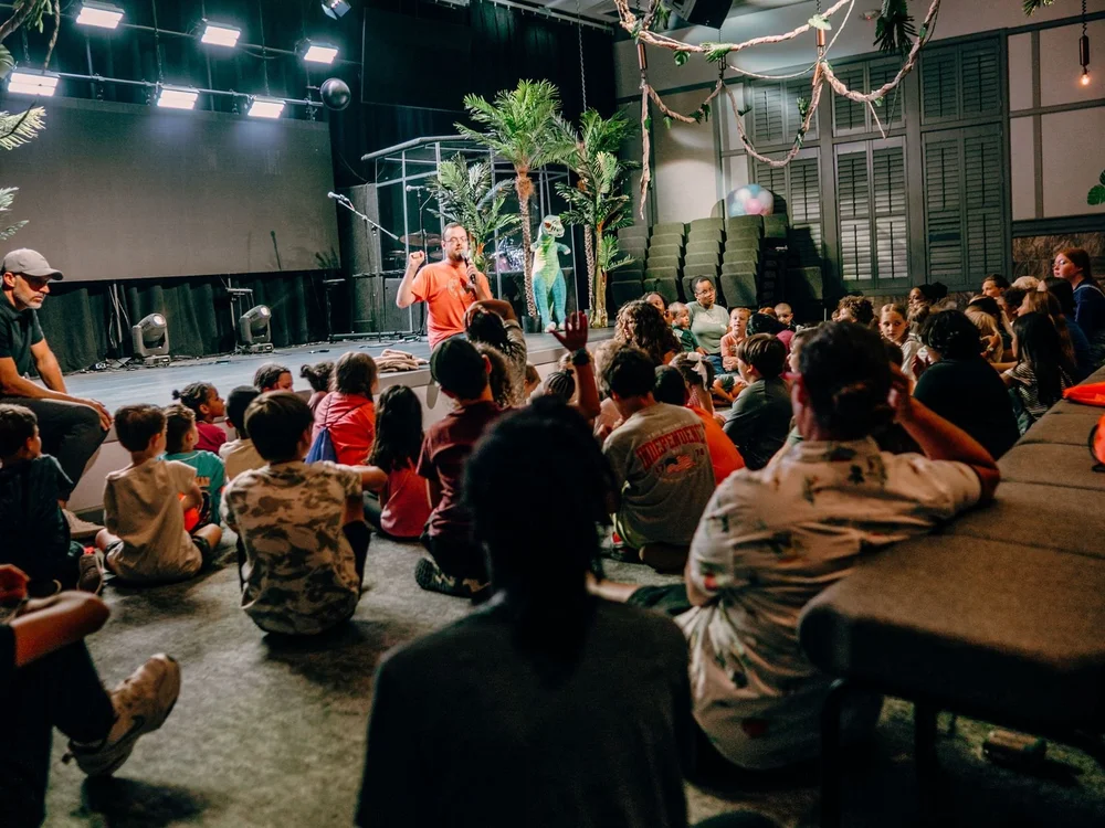 A room full of kids raising hands and listening to a leader