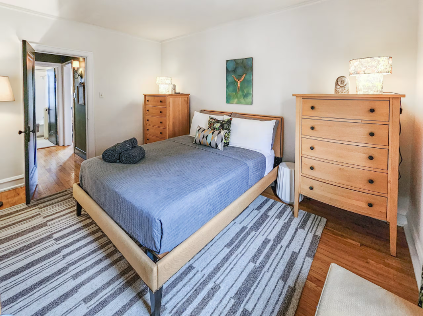 A Bedder Place: Bedroom with a gray-covered bed, wooden dressers, lamps and a striped rug.