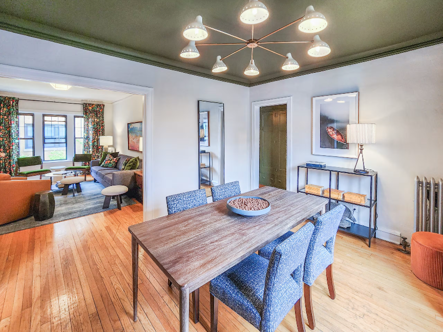 A Bedder Place: Dining room with a wooden table, patterned blue chairs and a branching ceiling fixture, opening into the living room.
