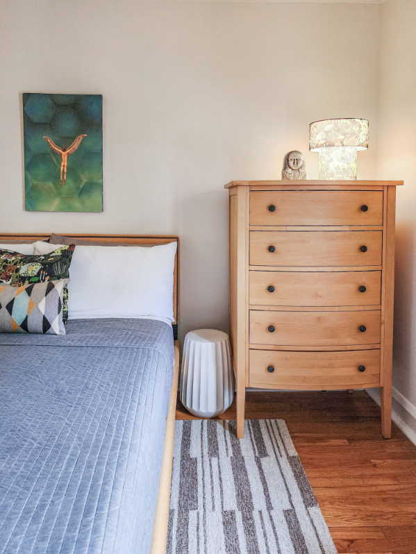 A Bedder Place: Portrait bedroom detail showing the bed, wooden dresser, lamp and wall artwork.