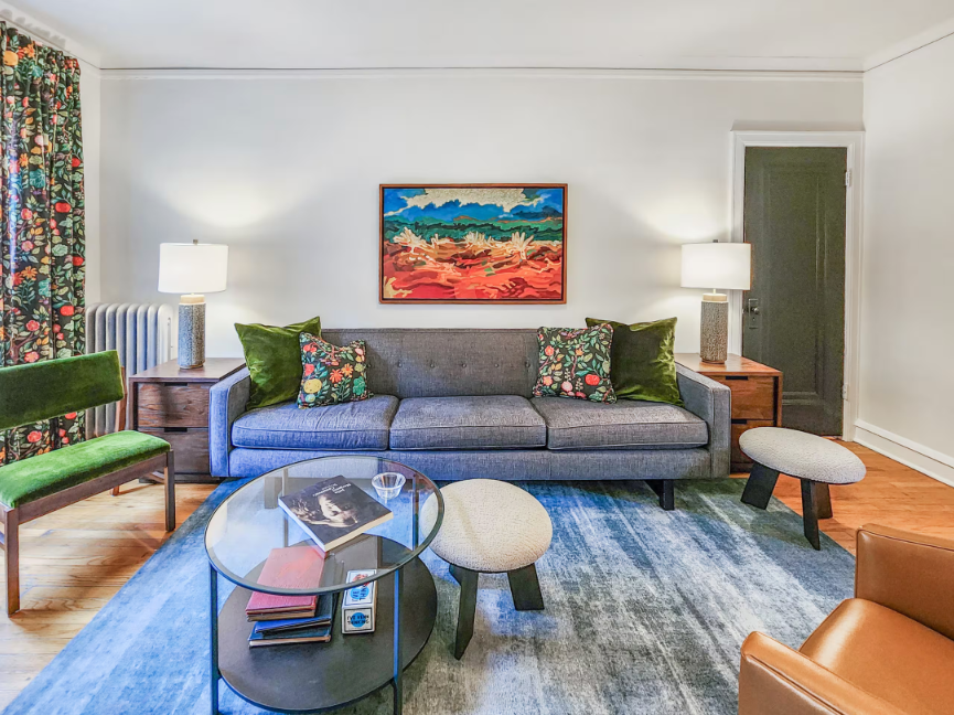 A Bedder Place: Frontal view of the gray living room sofa with green cushions, side tables, lamps and colorful artwork.