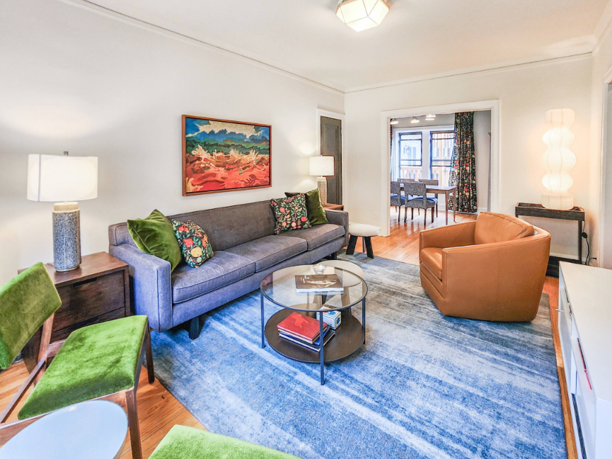 A Bedder Place: Living room with a gray sofa, green upholstered chairs, caramel armchair, glass coffee table and blue rug.