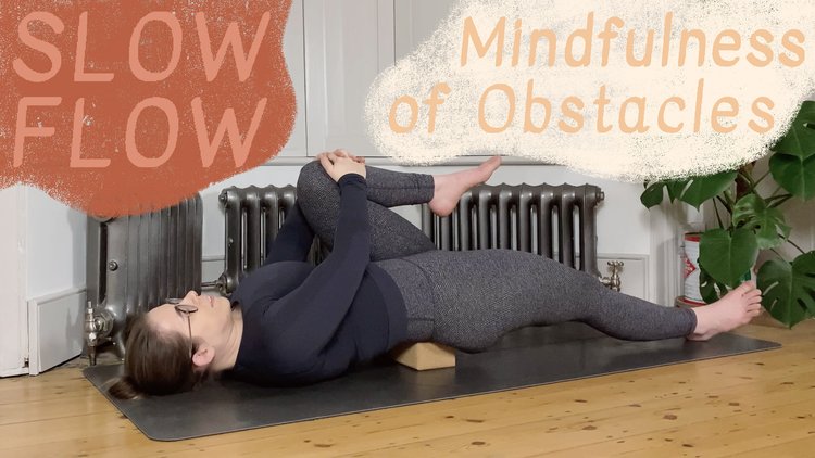 Oceana is lying on her yoga mat with a cork brick supporting her lower back, drawing one knee towards her chest with her hands interlaced.