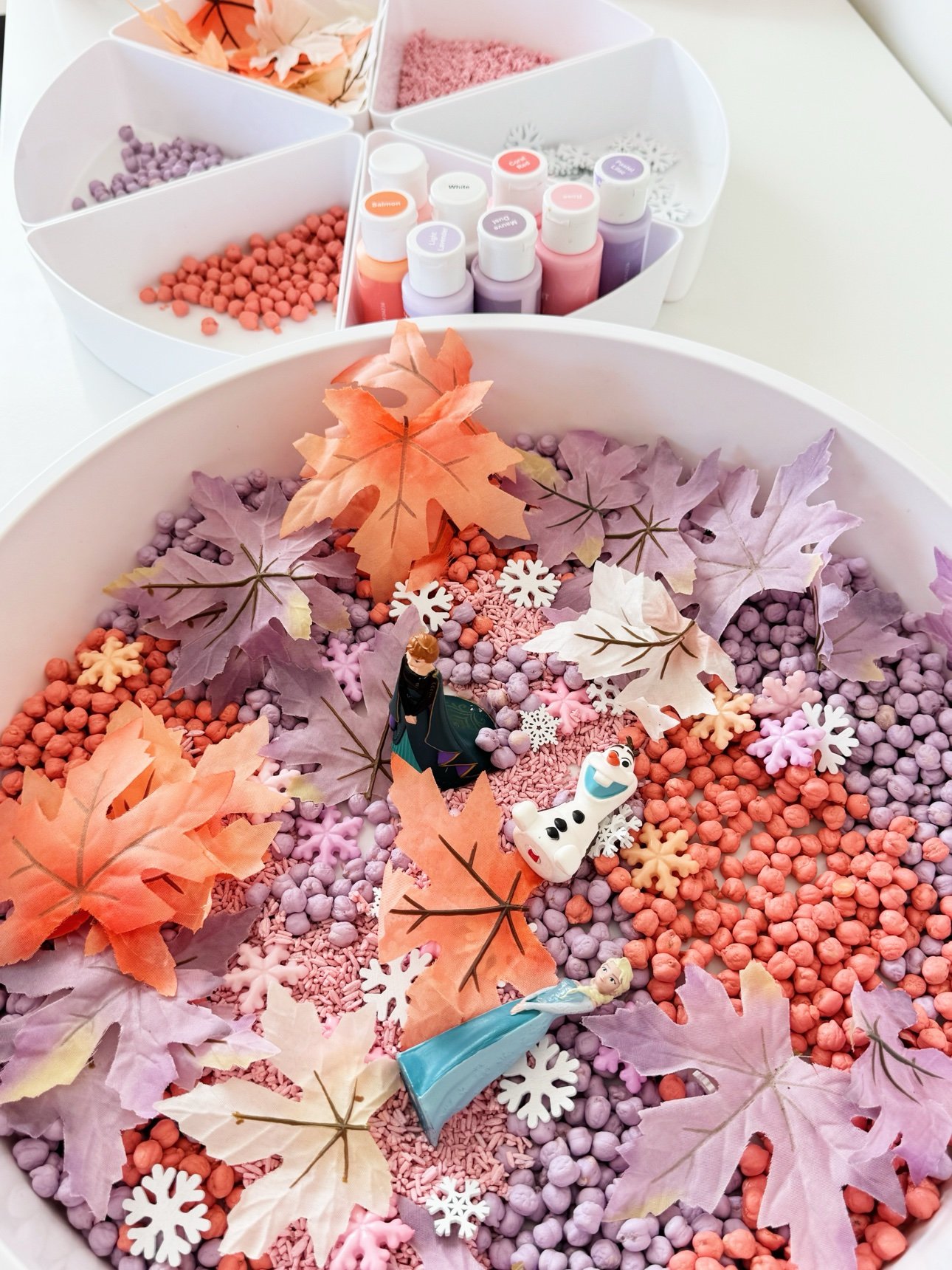 Finished fall frozen sensory bin with the segmented tray of extra dyed fillers, leaves, snowflakes, and paints behind it