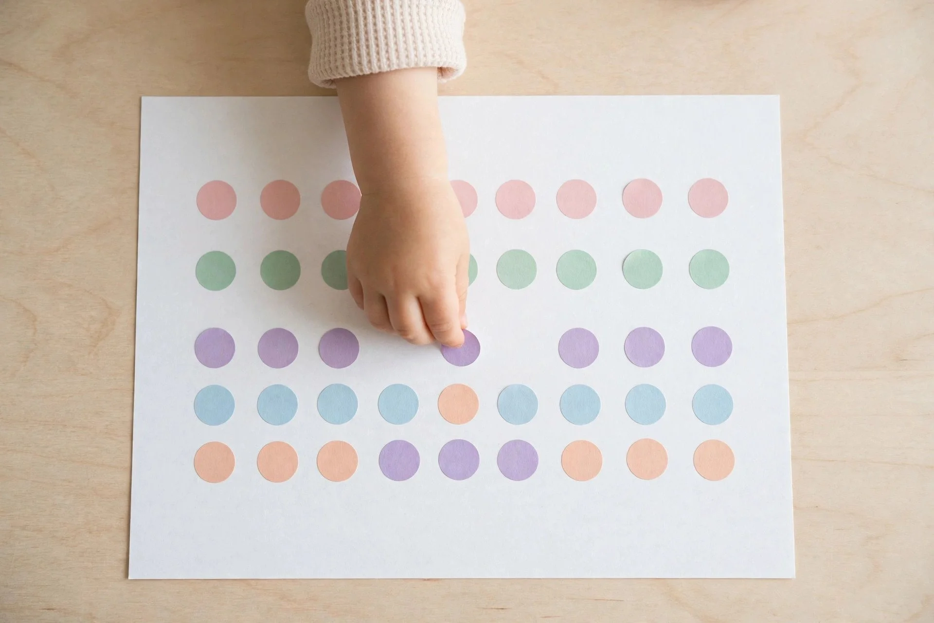 Toddler hand placing a pastel dot sticker onto plain white paper