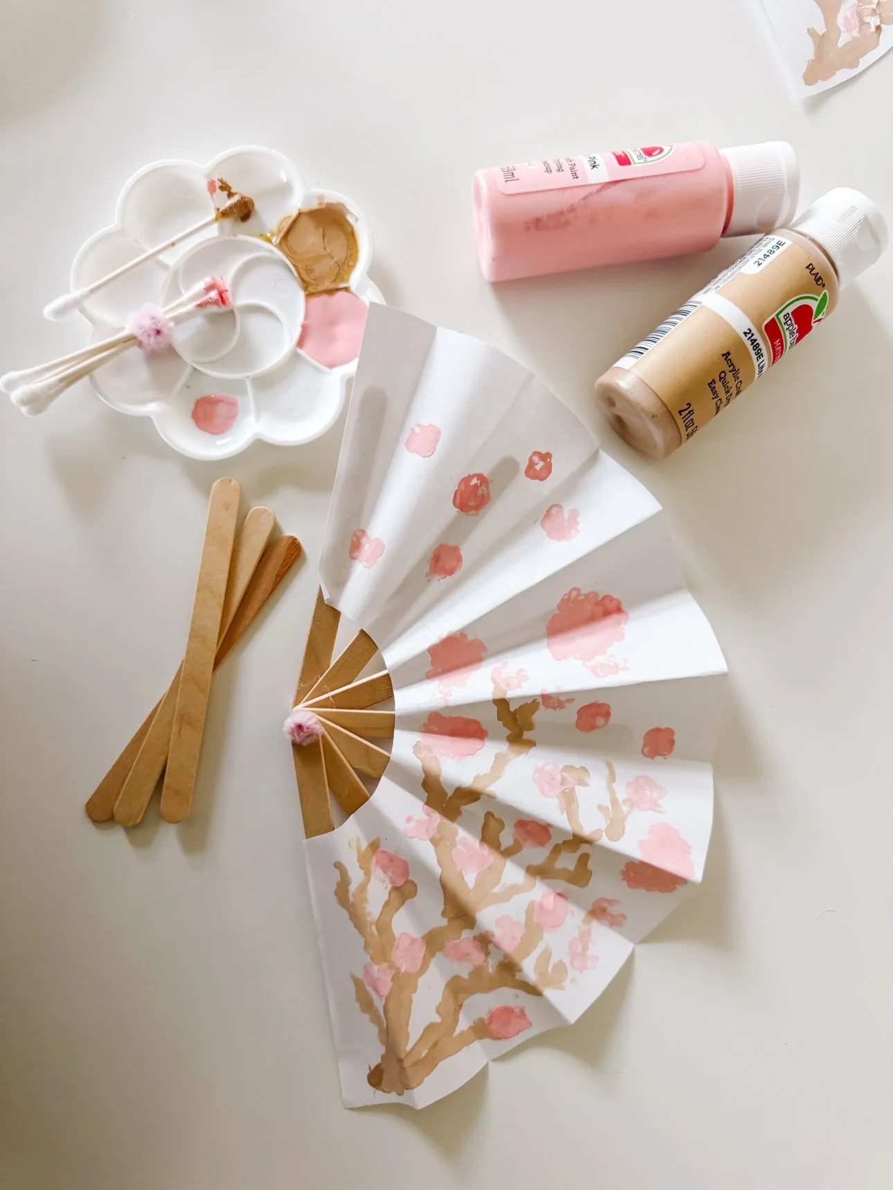 Cherry blossom paper fan being painted, with popsicle sticks, acrylic paint, and cotton swabs nearby