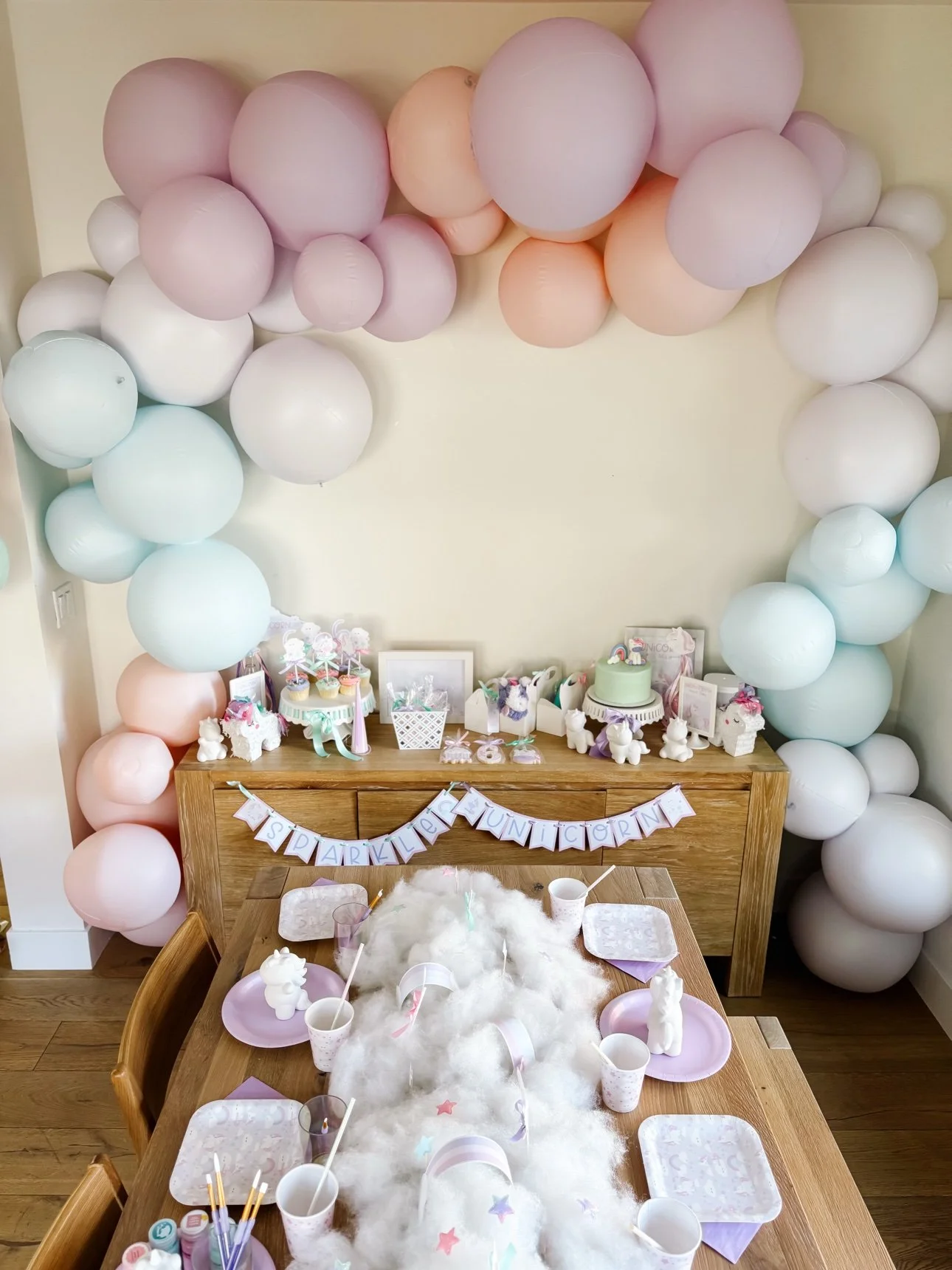 Unicorn party setup with pastel balloon arch, dessert table and kids table with cloud runner