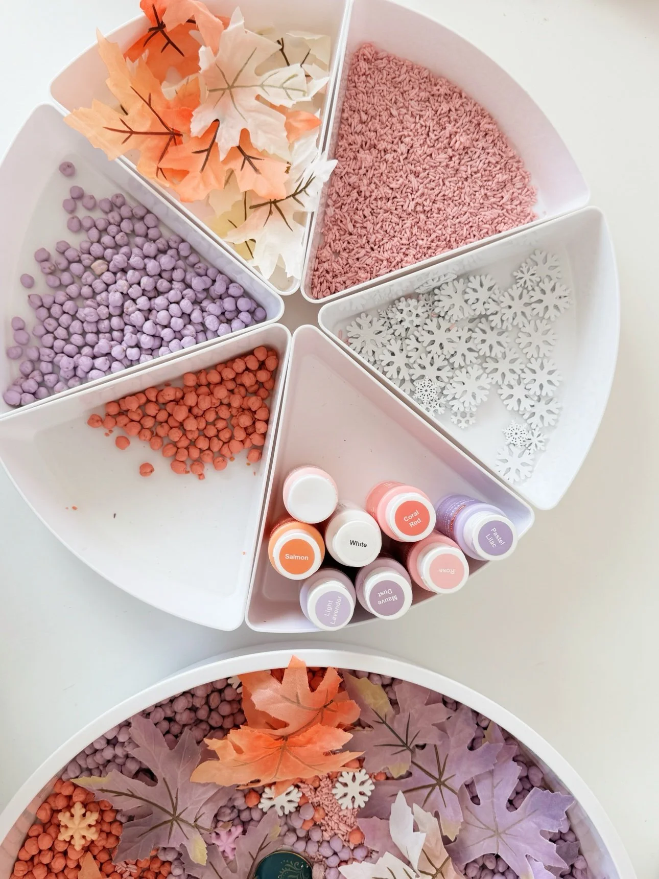 Fall frozen sensory bin ingredients in a round white segmented play tray: dyed lilac chickpeas, coral chickpeas, blush pink rice, pastel fall leaves, white wooden snowflakes, and pastel acrylic paint bottles