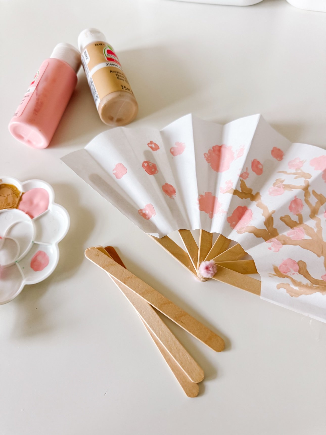 Painted cherry blossom paper fan lying flat with pink and tan paint bottles and popsicle sticks