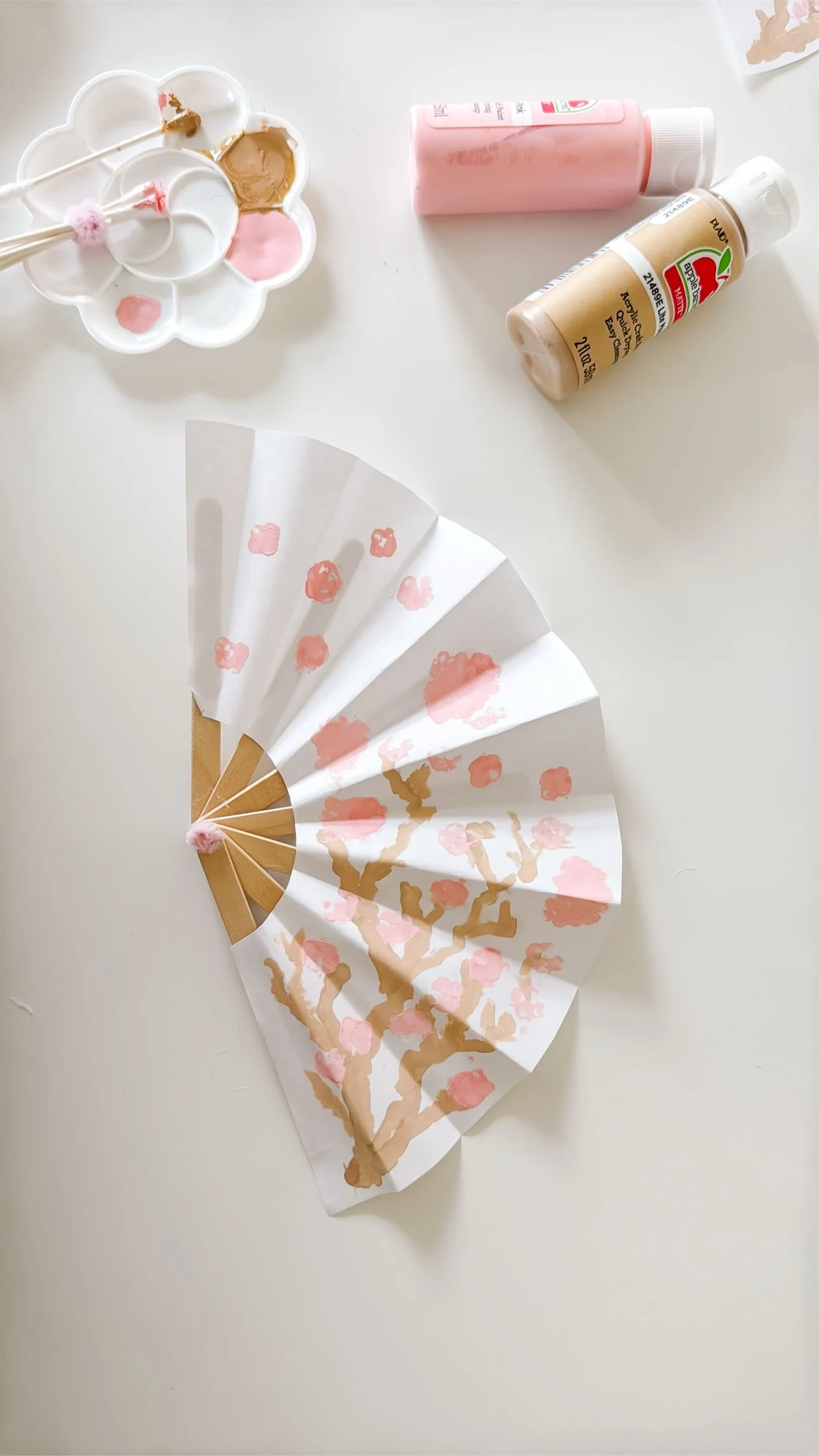 Finished cherry blossom paper fan with pink and tan acrylic paint bottles nearby