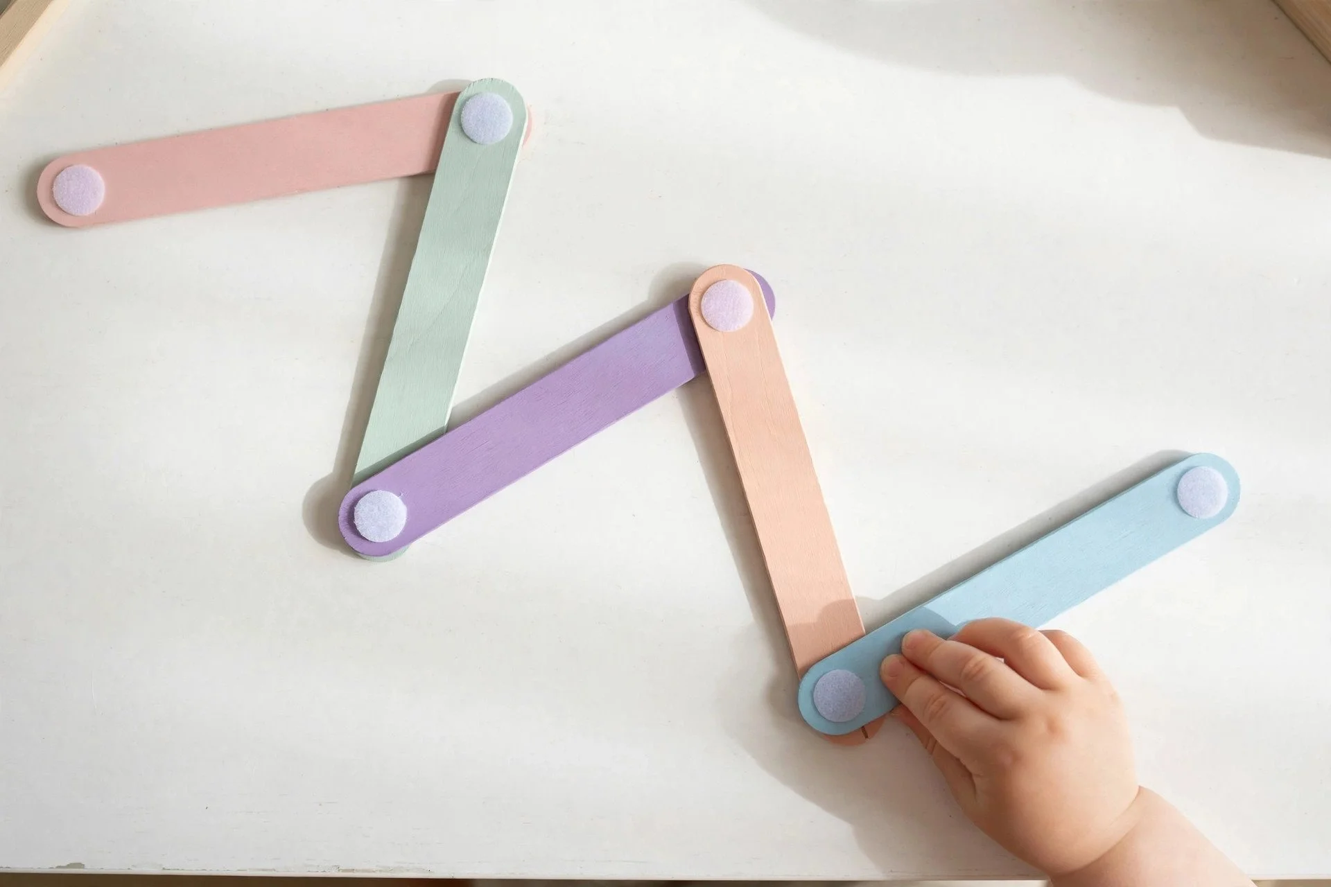 Pastel popsicle sticks with velcro dots snapped together in zigzag chains, toddler hand connecting them