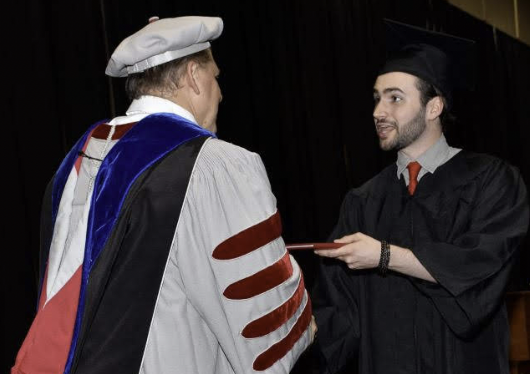 A graduate in cap and gown shakes hands with a university official in academic regalia while receiving a diploma cover