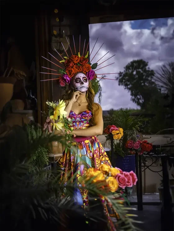 Catrina in a vivid floral dress and red rose headdress posing beside a window