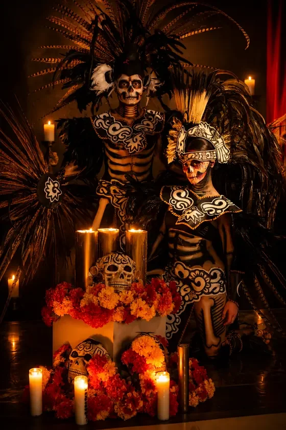 Two Catrina participants in black feathered costumes posing by candles and marigolds