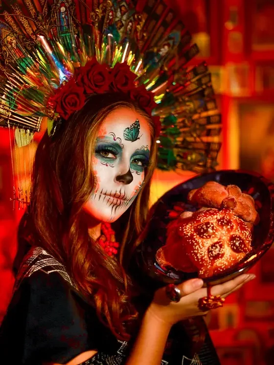 Catrina with a radiant red headdress holding decorated pan de muerto