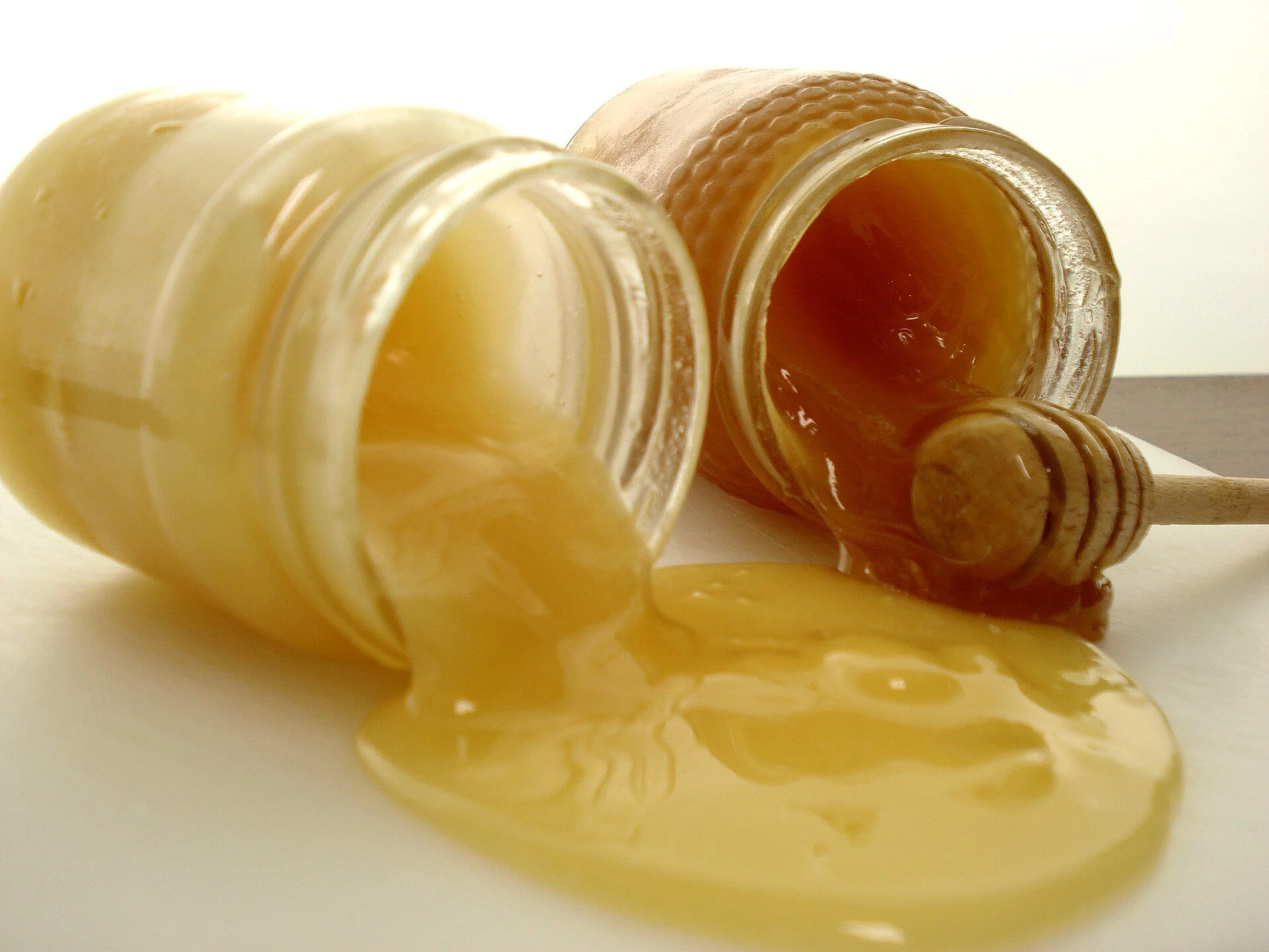 Two jars of honey tipped over, honey pooling on a white surface with a wooden dipper