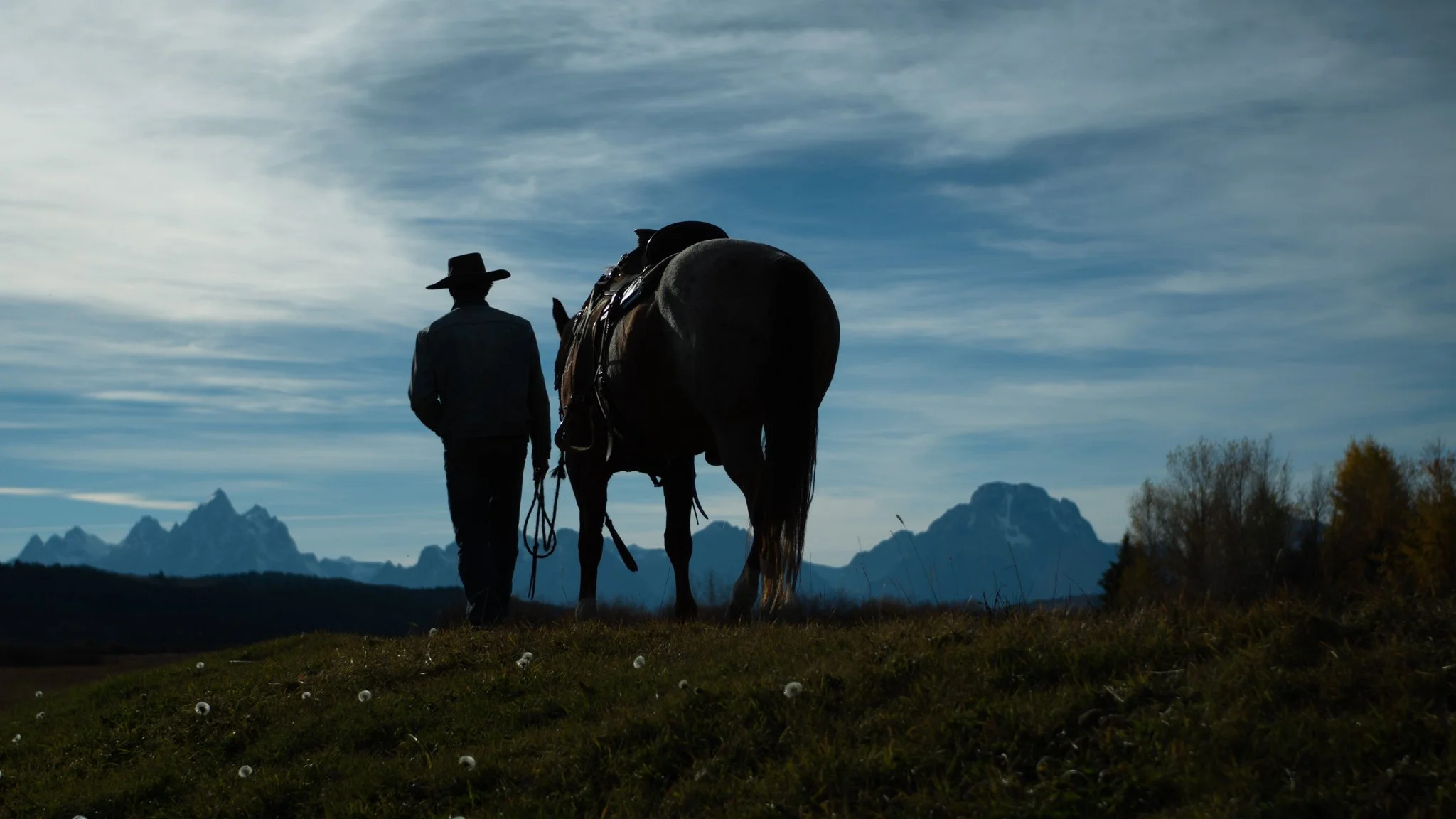 A cowboy and saddled horse silhouetted against the Tetons