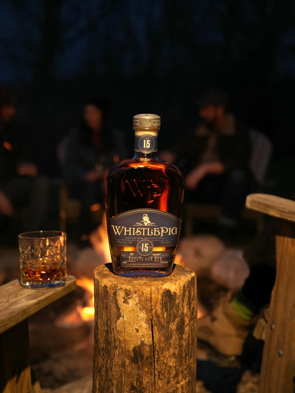 WhistlePig bottle and glass on wooden stumps beside a fire