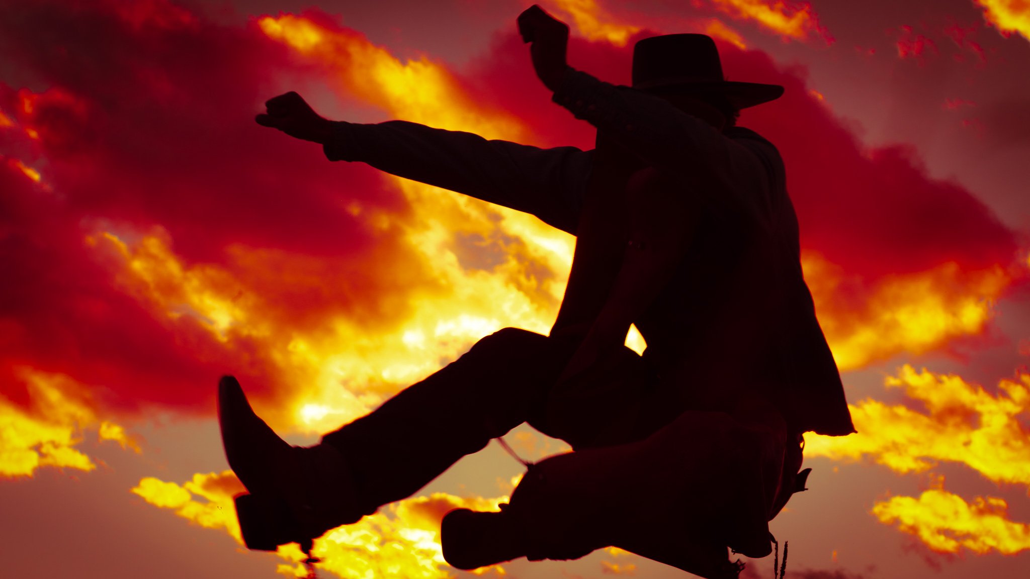 A cowboy silhouetted in midair against fiery clouds