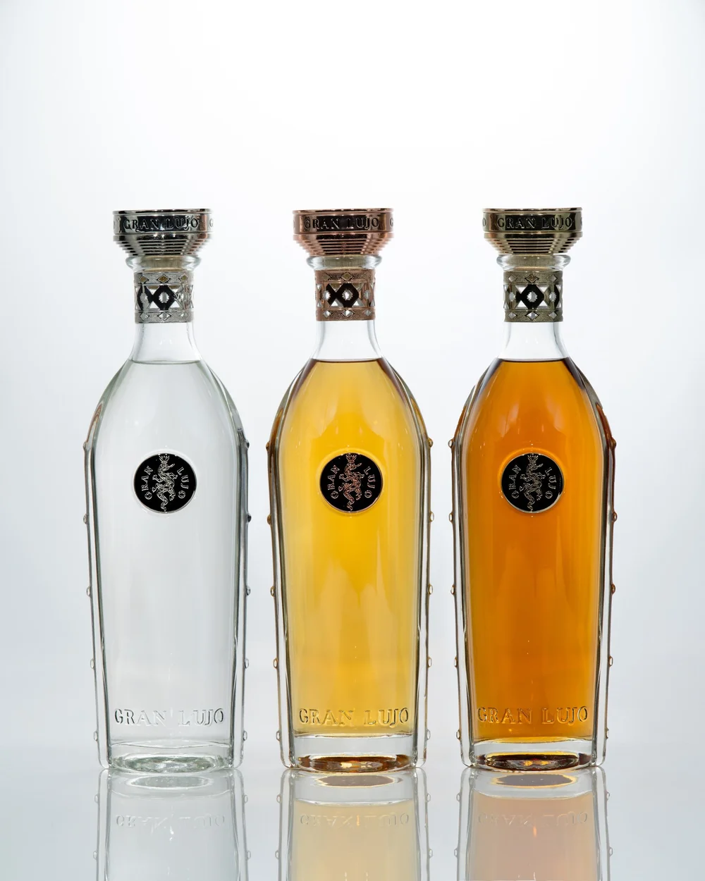 Three Gran Lujo bottles on a reflective surface against a white background