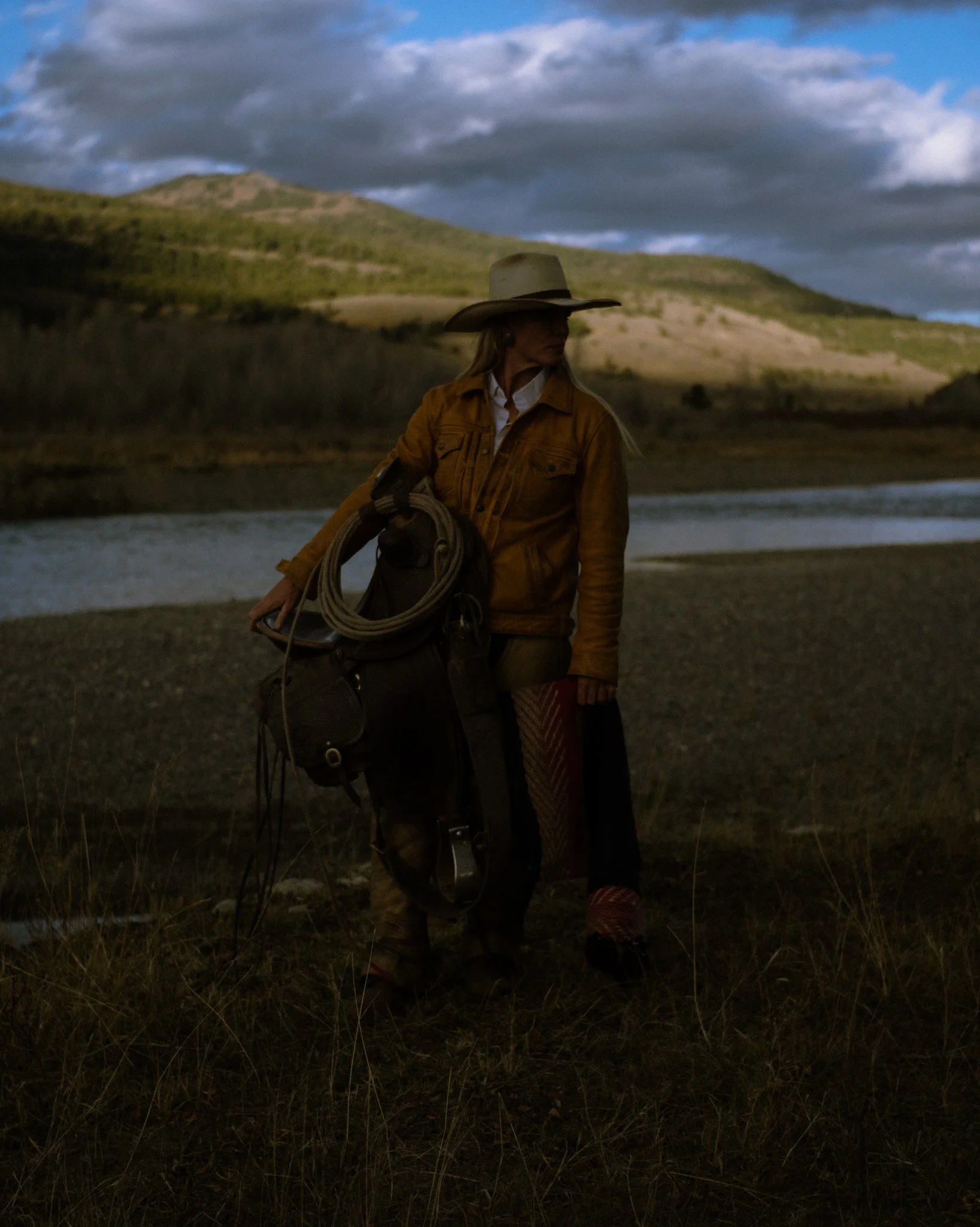 A cowgirl holding a saddle beside a river