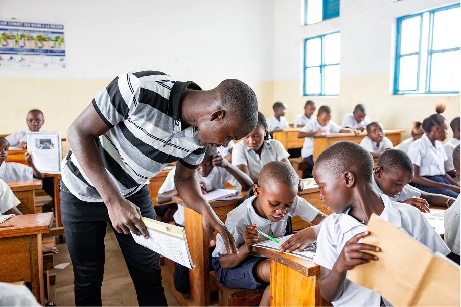Teacher leans over a student’s desk, guiding children as they work on written assignments in a crowded classroom.