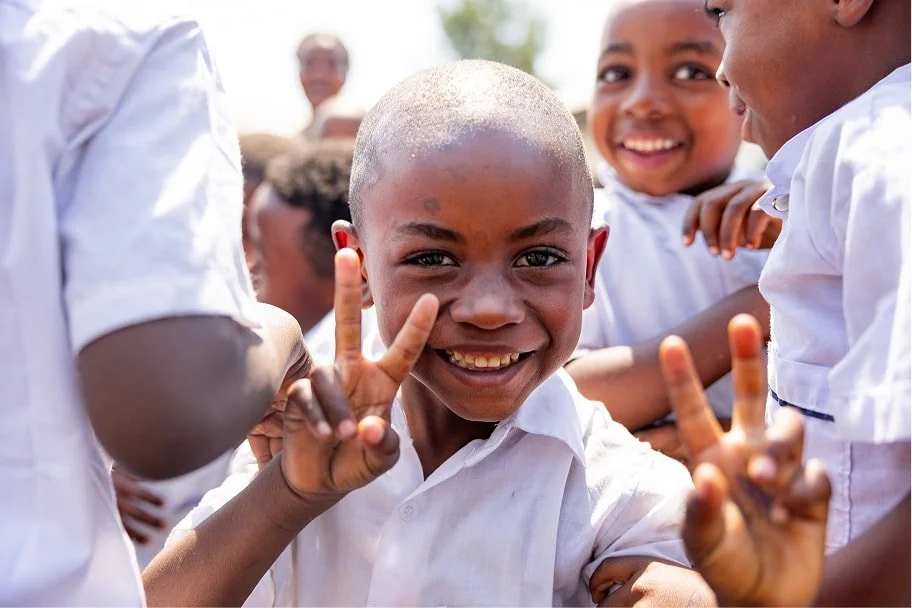 A smiling student holds up two peace signs while standing with classmates outdoors at school.