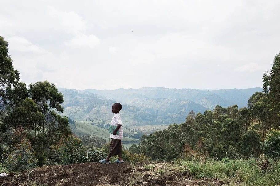 A young child stands on a hillside overlooking a green valley and distant mountains.