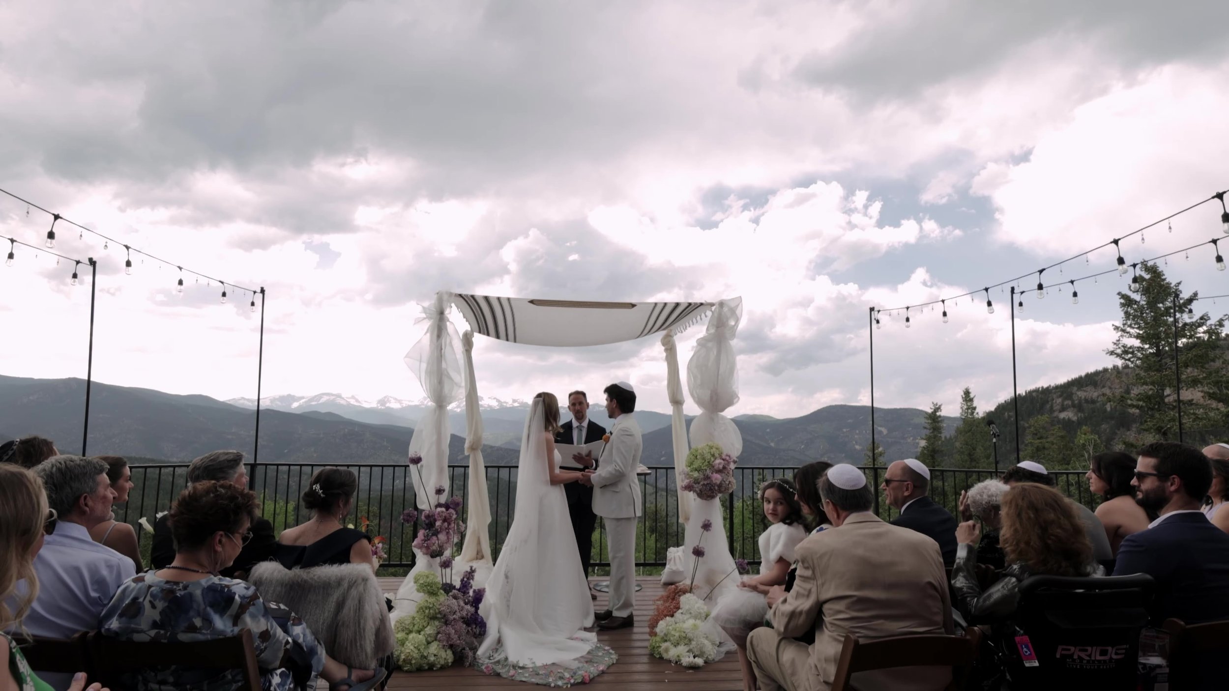 Wide outdoor ceremony under a white chuppah on the deck at Northstar Gatherings in Idaho Springs, Colorado, mountains behind the guests
