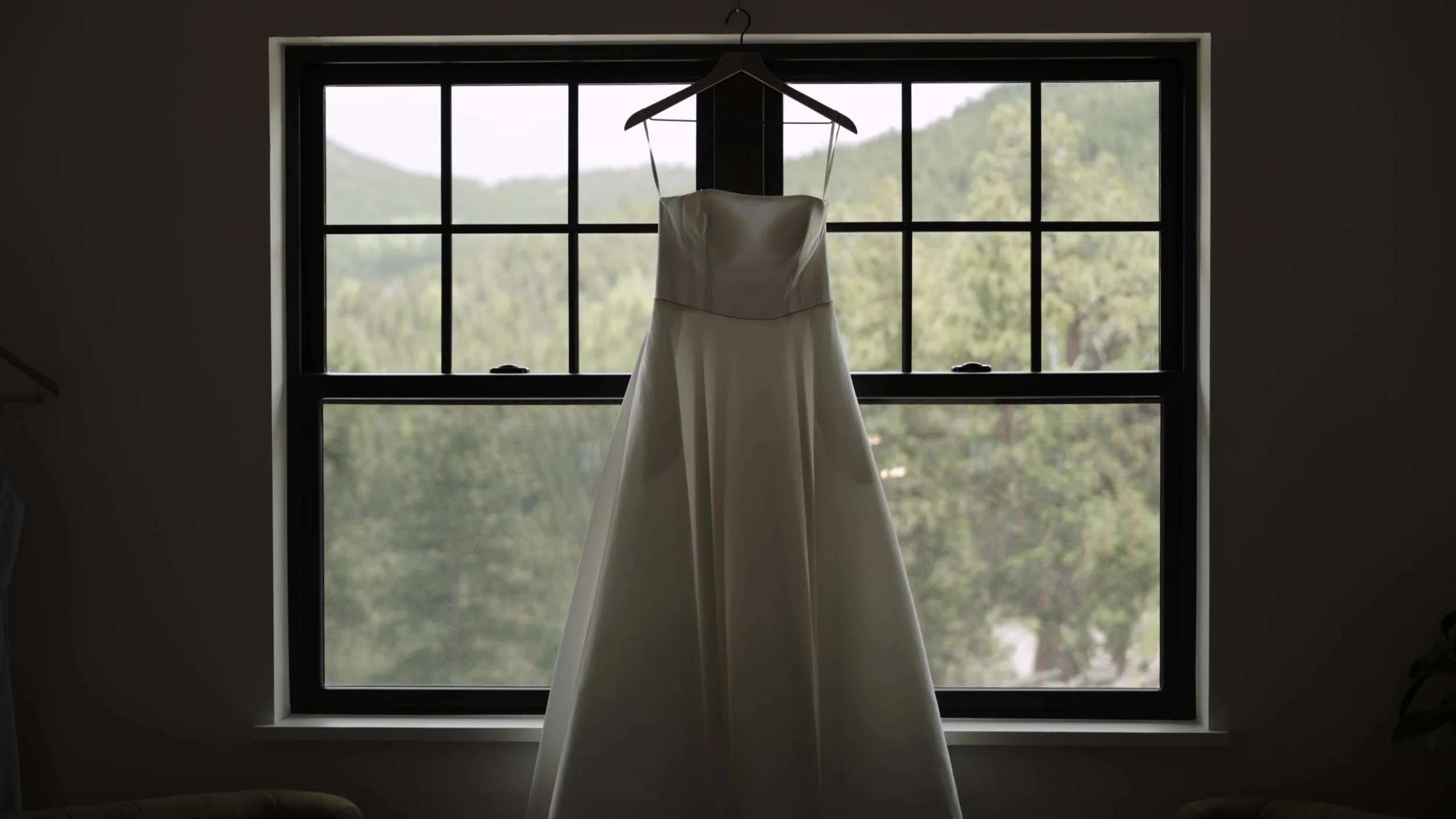 White wedding dress hanging in a black-framed window overlooking pine forest at Northstar Gatherings in Idaho Springs, Colorado