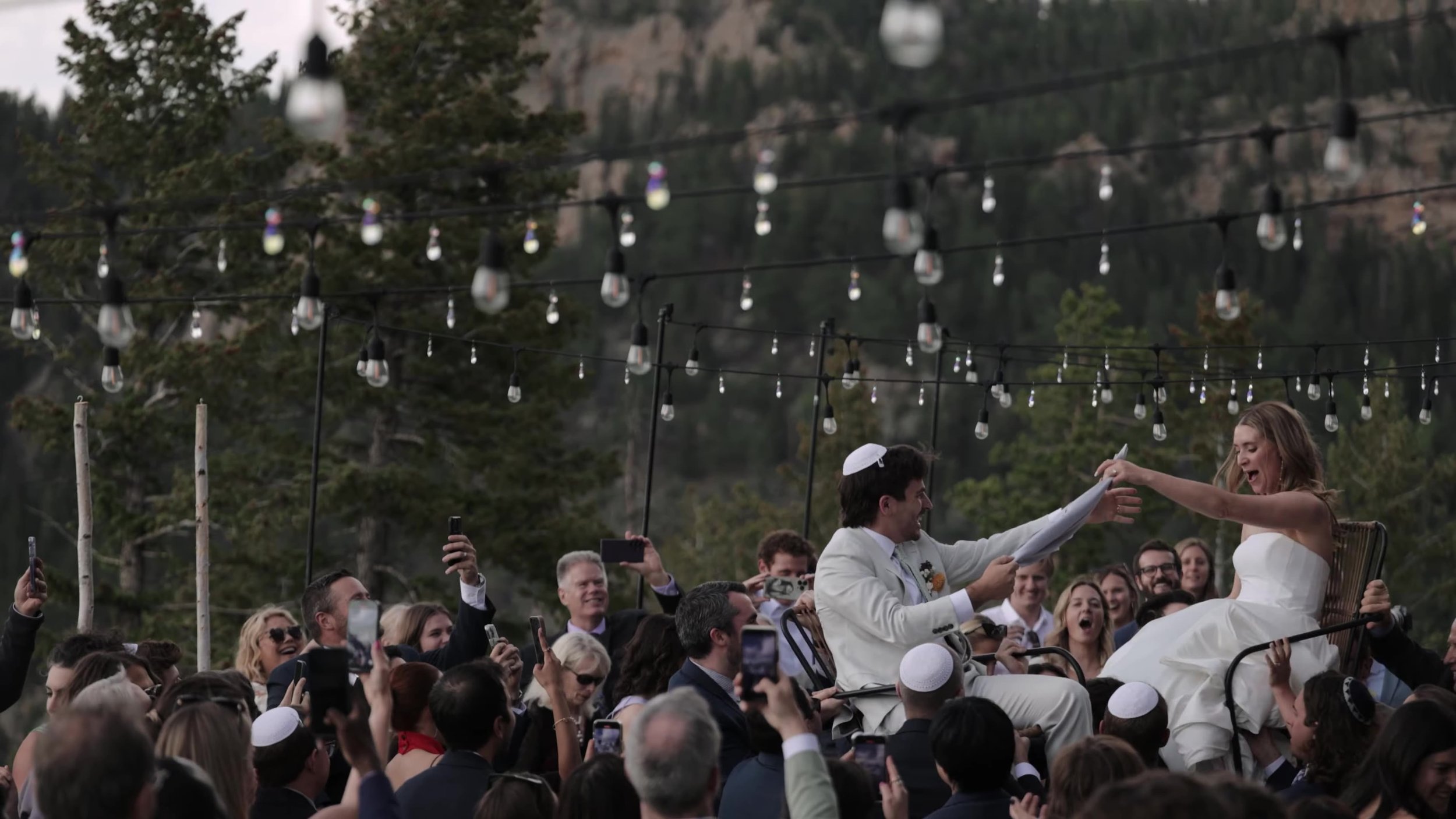 Bride and groom lifted on chairs during the hora under string lights at Northstar Gatherings in Idaho Springs, Colorado
