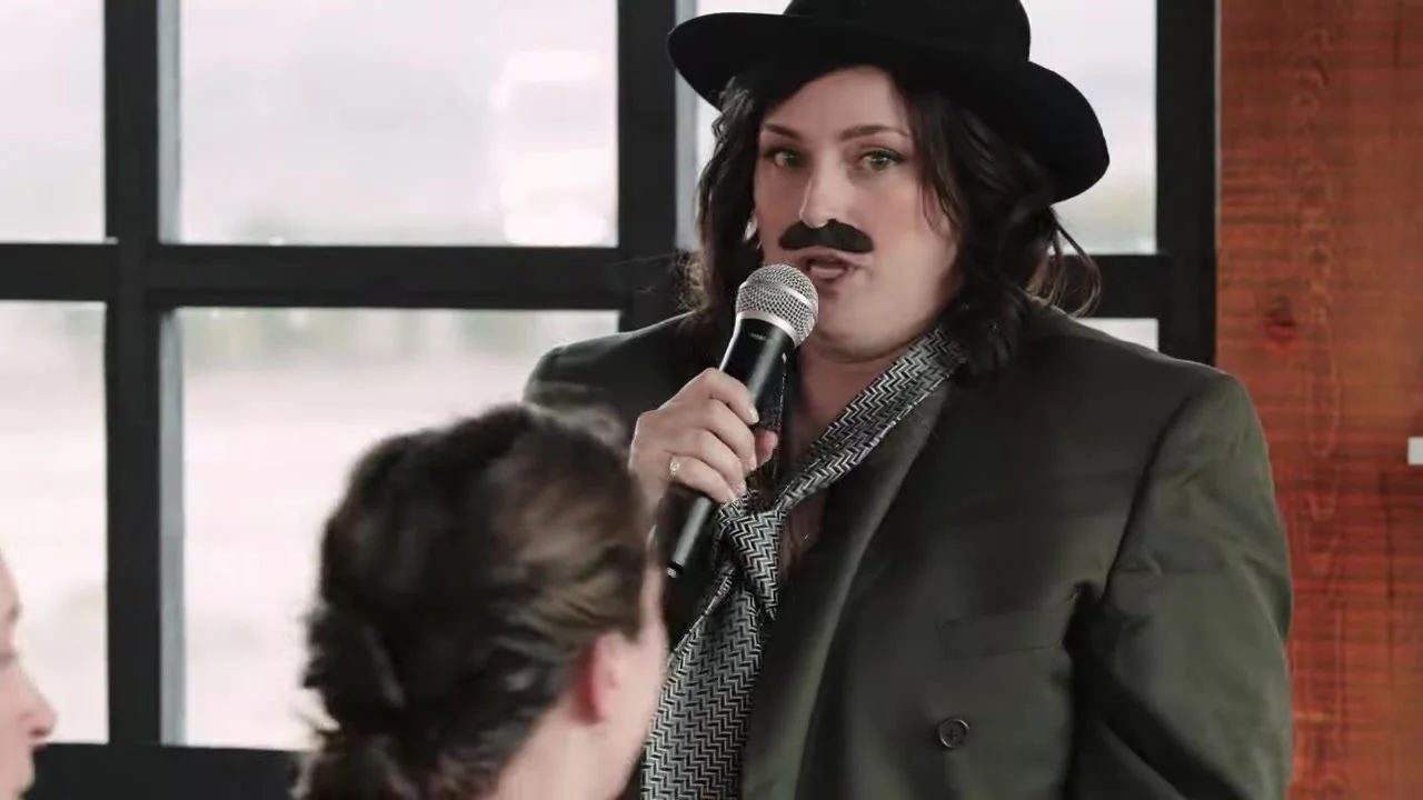 Guest in a black fedora and fake handlebar mustache speaking into a silver microphone at 3 Peaks Ranch