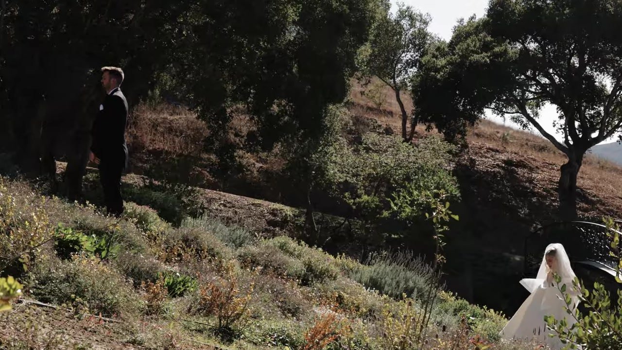 A groom in black waits under an oak while the bride in a long veil walks up a dry hillside