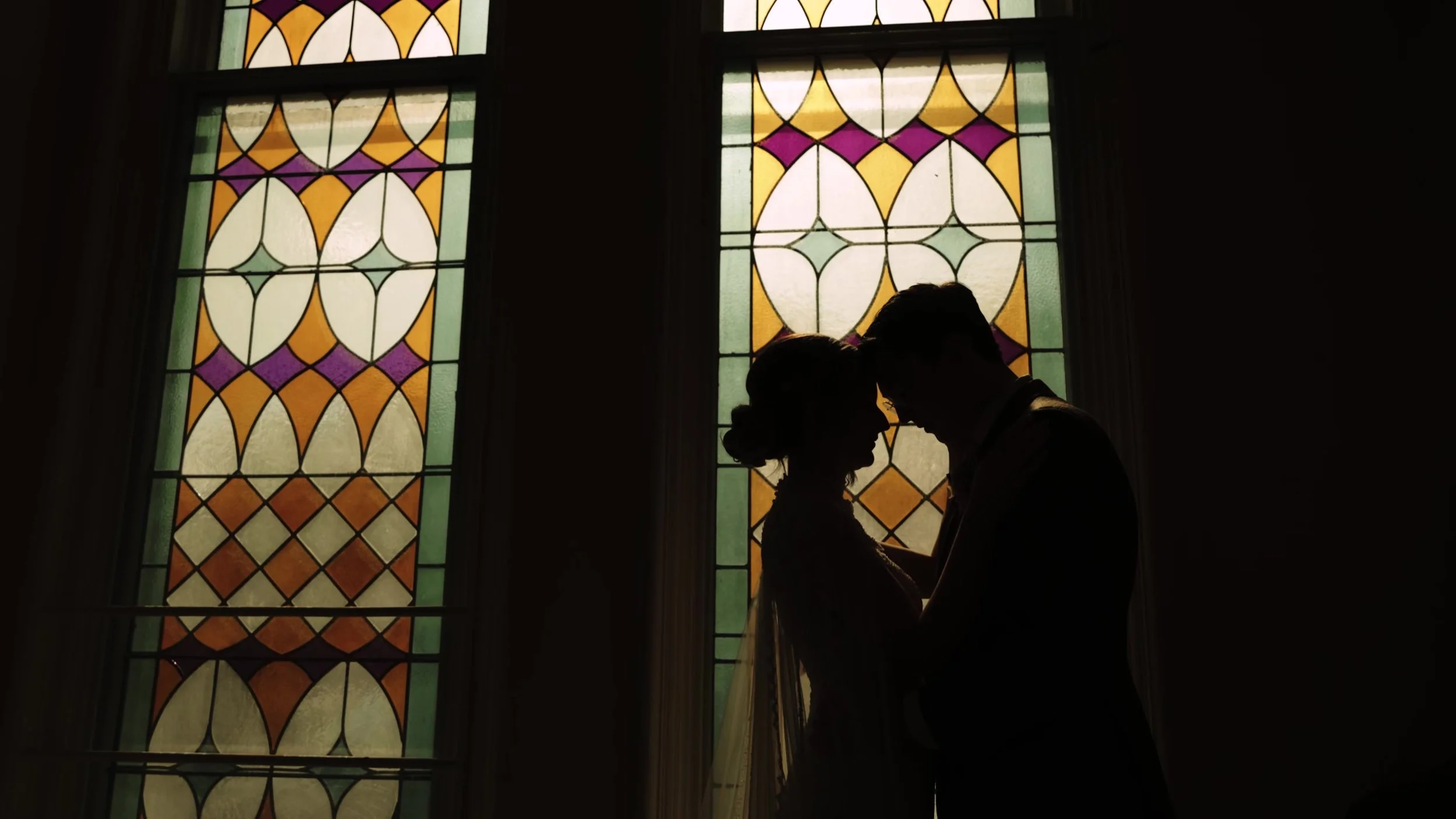 Silhouette of bride and groom forehead to forehead between stained glass windows at Nouveau Antique Art Venue in Houston