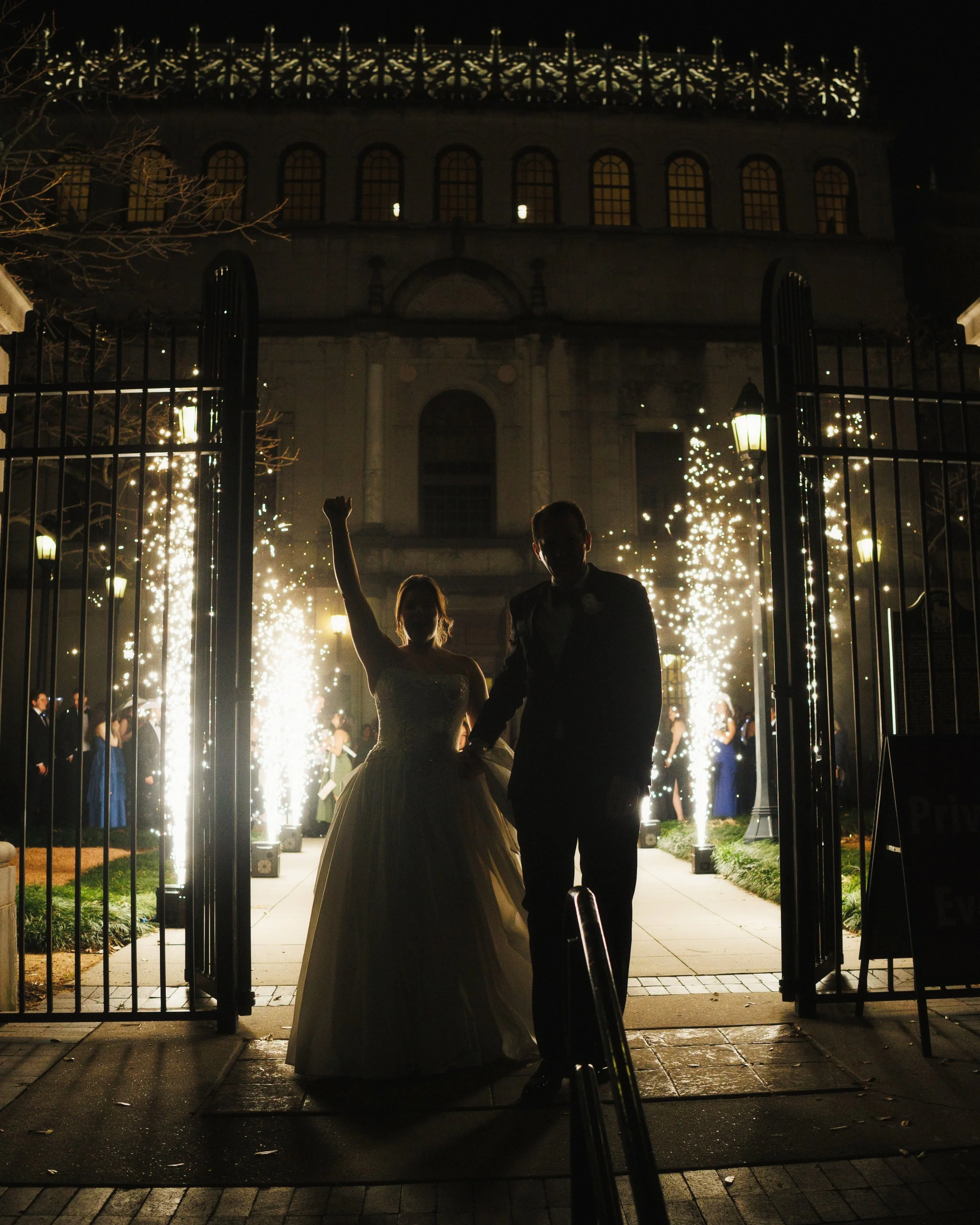 Bride and groom celebrating during a sparkler send-off through an iron gate at night