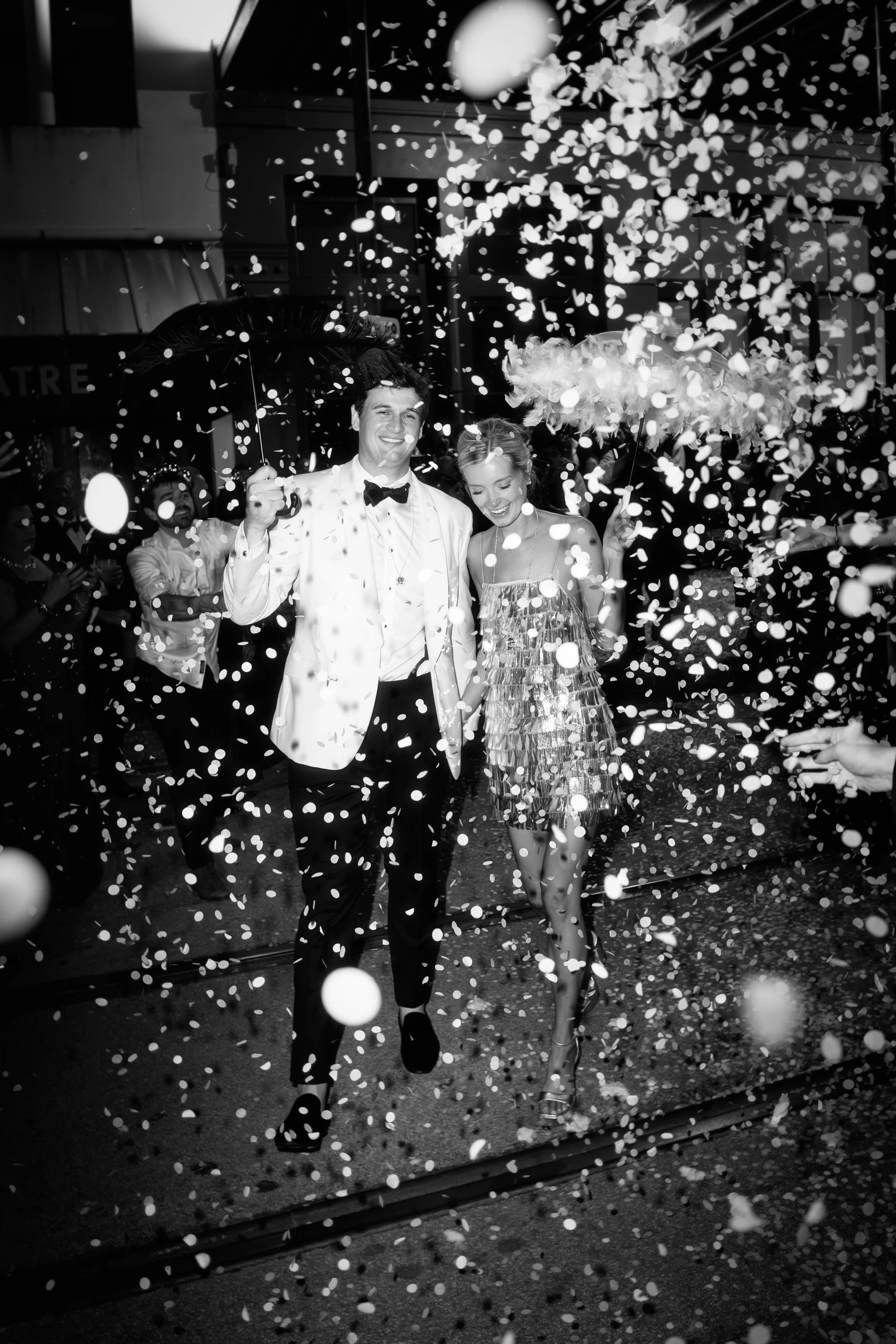 Black and white photo of bride and groom walking through a shower of confetti holding feathered parasols