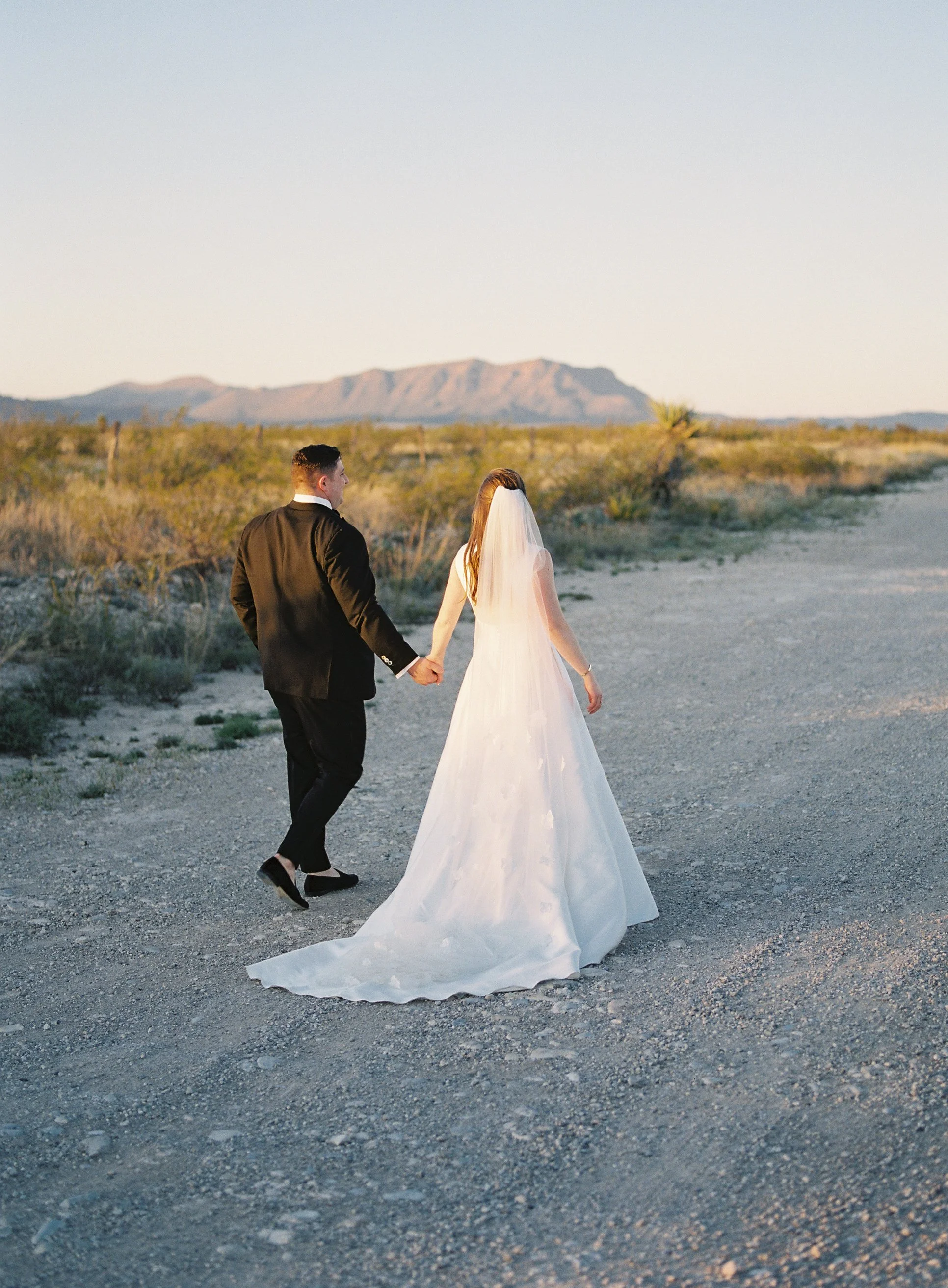 Bride and groom walking hand in hand down a desert road at golden hour with mountains beyond