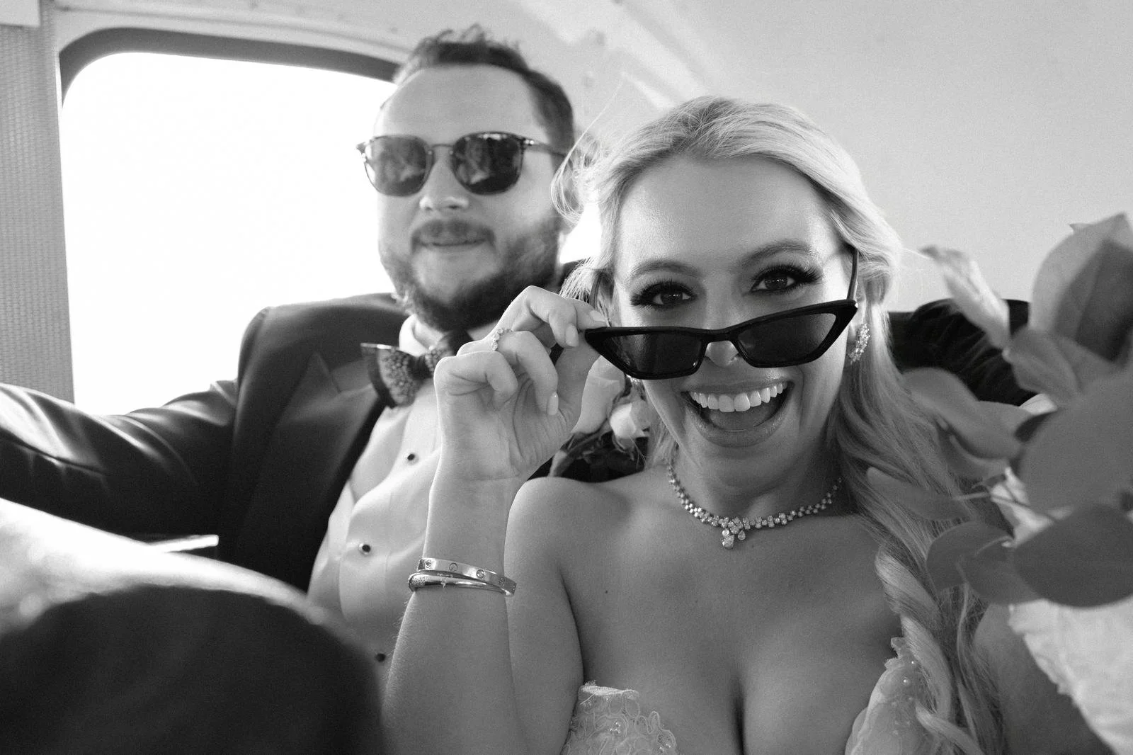 Black and white close up of bride and groom laughing in sunglasses inside a car