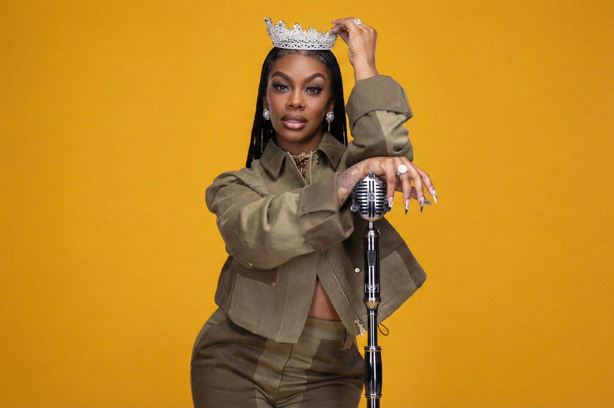 Jess Hilarious wearing a crown, standing at a microphone against a gold backdrop
